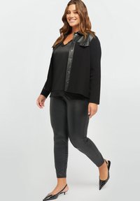 Zwarte getailleerde leggings, satijnen camisole en een zwarte jas met leren accenten. Het model draagt puntige zwarte schoenen met witte stippen.