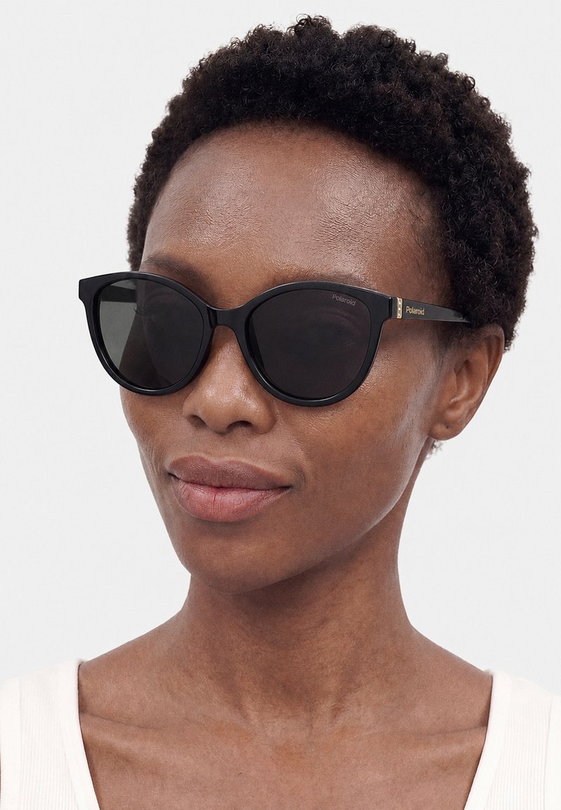 Woman with short curly hair wearing black cat-eye sunglasses and a white sleeveless top against a plain background.