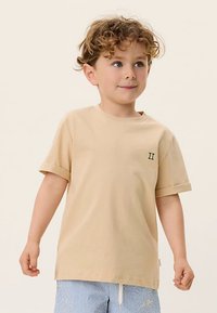 Jaunas garbanotas plaukų berniukas, dėvintis smėlio spalvos trumpomis rankovėmis marškinius su sulankstytomis rankovėmis ir mėlynos dryžuotos šortus, stovintis prieš paprastą foną.