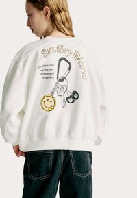 Sweatshirt en coton blanc avec une coupe oversize, arborant une impression graphique de clés et d'un smiley au dos. Poignets et taille côtelés.