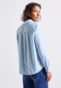 Vrouw met kort krullend haar, die een lichtblauw gestreept overhemd met lange mouwen en blauwe jeans draagt, kijkt over haar schouder tegen een witte achtergrond.