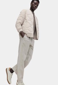 Beige kviltad bomberjacka med ribbade mudd, kombinerad med ljusgrå byxor i avslappnad passform och vita sneakers med bruna detaljer.