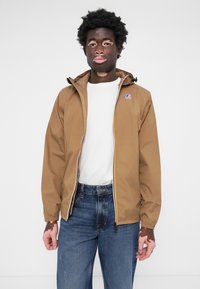 Brown hooded jacket with a zip front, yellow accent, and logo patch. Paired with a white shirt and blue jeans. Smooth, lightweight texture.