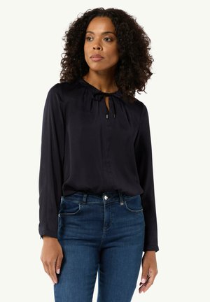 Navyblaue Seidenbluse mit einem Ausschnitt in Form eines Schlüssellochs, langen Ärmeln und subtilen Falten am Kragen. Kombiniert mit dunkelblauen Skinny-Jeans.
