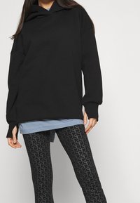 Svart stor hoodie med huva, långa ärmar och ribbade muddar, lager på lager över en ljusblå skjorta och mönstrade svarta leggings.