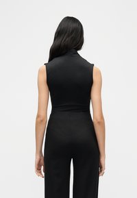 Γυναίκα με μακριά σκούρα μαλλιά, ντυμένη με αμάνικο μαύρο τοπ ζιβάγκο και μαύρο παντελόνι με ψηλή μέση, στέκεται αντικρίζοντας ένα λευκό φόντο.