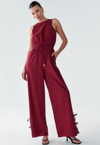 Burgundski kombinezon z zategnjenim pasom, širokimi hlačnicami in pentljami na spodnjih robovih. Iz mehkega blaga, brezrokaven dizajn.