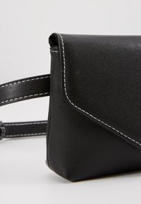 Černá kabelka z umělé kůže s klopou a bílým prošíváním. Má štíhlý tvar a nastavitelný popruh pro všestranné používání.