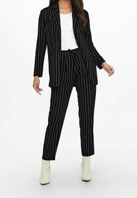 Černý kostým s pruhy, který obsahuje dlouhý blazer a zúžené kalhoty, kombinovaný s bílým topem a bílými lodičkami na kotníky.
