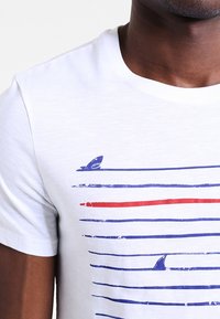 Bílá tričko s modrými vodorovnými pruhy a červeným pruhem, s žraločími ploutvemi, které se objevují poblíž oblasti hrudníku.