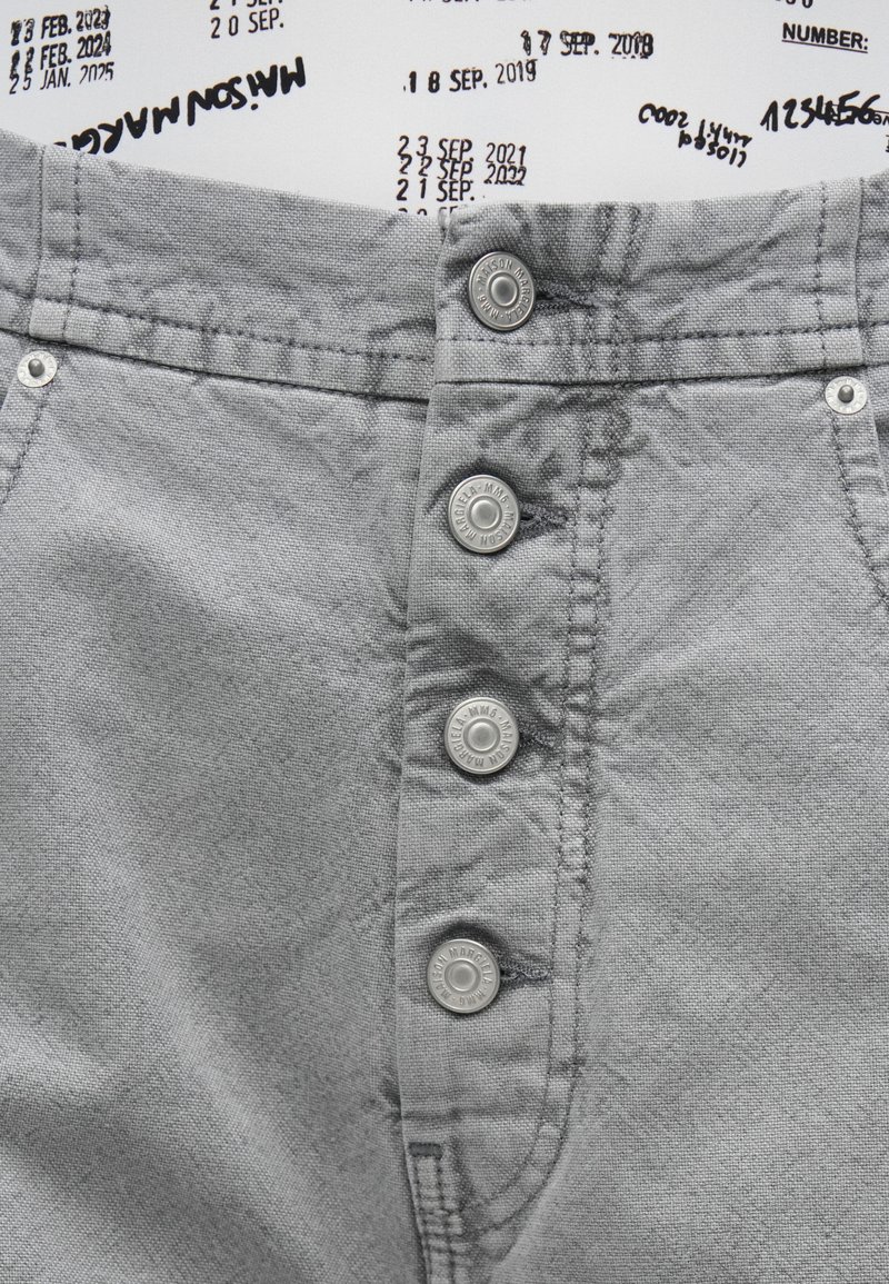 Graue Stoffshorts mit hoher Taille, fünf Knöpfen und Metallakzenten. Die Textur wirkt weich mit einem leicht zerknitterten Finish.