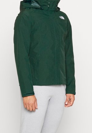 Blouson à capuche vert foncé avec fermeture éclair à l'avant et poignets en velcro, porté avec un pantalon gris clair, montrant le haut du corps et les bras.