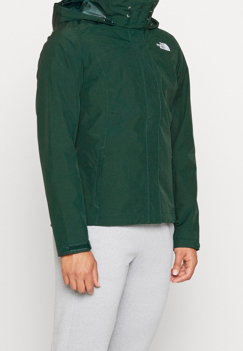 Blouson à capuche vert foncé avec fermeture éclair à l'avant et poignets en velcro, porté avec un pantalon gris clair, montrant le haut du corps et les bras.