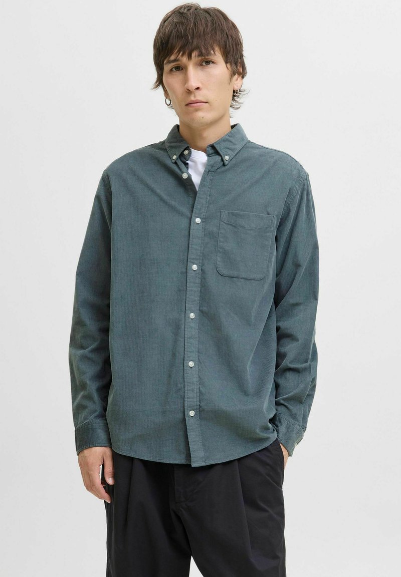 Mørkegrønn skjorte med knapper og lange ermer, med en brystlomme og teksturert stoff. Brukt over en hvit t-skjorte.