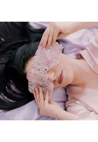 Różowa, teksturowana maska na oczy wykonana z plecionego materiału, trzymana w dłoniach, na gładkim tle przypominającym satynę.