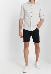 Camisa de lino beige claro con mangas remangadas y un bolsillo frontal, combinada con pantalones cortos negros y zapatillas blancas. Diseño minimalista y corte slim.