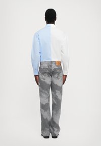 Chemise bleu clair et blanche à demi-boutonnage, associée à un jean gris avec un motif nuage et une étiquette en cuir marron sur la taille.
