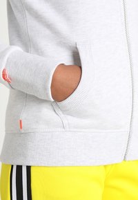Felpa grigia con zip e polsini e fondo a costine, dotata di una tasca laterale con un accento arancione. Abbinata a pantaloni gialli con strisce nere.