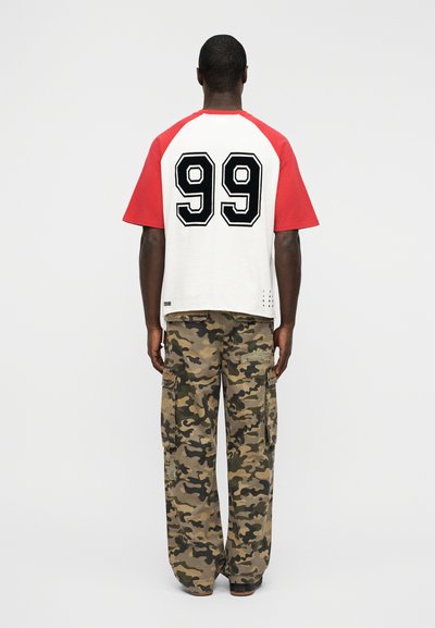 Κοντομάνικο ράγκλαν μπλουζάκι σε κόκκινο και άσπρο με μαύρο αριθμό "99" τυπωμένο στην πλάτη, συνδυασμένο με φαρδιά παντελόνια καμουφλάζ τύπου cargo.