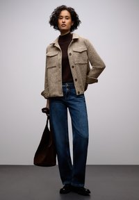 Béžová texturovaná bunda se dvěma náprsními kapsami přes tmavě hnědý rolák, doplněná volnými modrými džínami a hnědou kabelkou přes rameno.