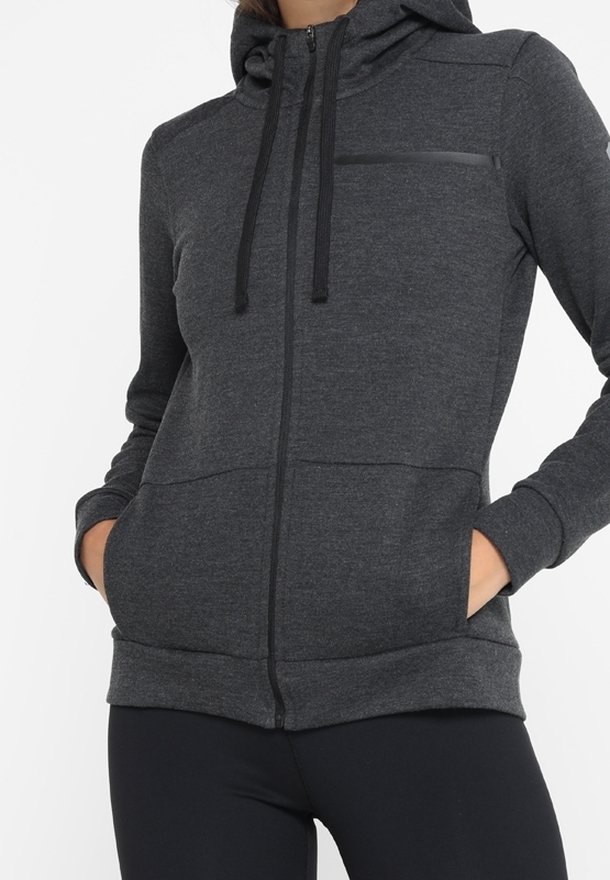 Sweat à capuche zippé gris foncé avec poches avant et cordons noirs, porté avec des leggings noirs, mains dans les poches, coupe décontractée avec capuche relevée.