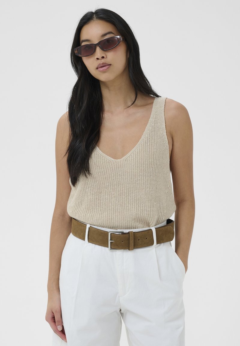 Femme aux longs cheveux noirs portant des lunettes de soleil foncées, un top beige en maille sans manches, un pantalon blanc taille haute et une ceinture en daim marron.