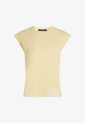 Be rankovių, šviesiai geltonos spalvos palaidinė su paminkštintais pečiais ir baltu kursyvu užrašu šalia apatinio krašto dešinėje pusėje.