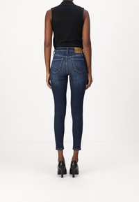 Vaqueros ajustados azul oscuro con una cintura de altura media, bolsillos traseros y un parche de cuero marrón claro en la cinturilla, combinados con tacones altos negros.