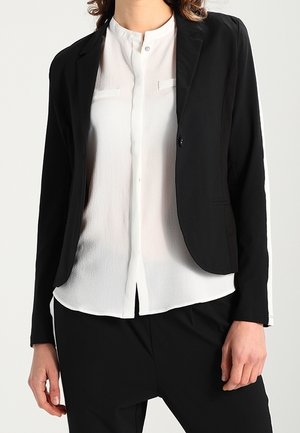 Vrouw draagt een wit gebuttonned bloesje met een zwart enkeknoopblazer en zwarte broek, getoond van nek tot heupen.