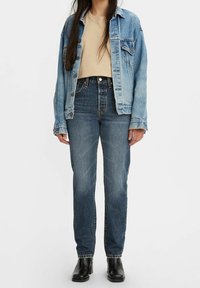 Chaqueta de denim en azul claro con áreas descoloridas, combinada con pantalones vaqueros de tiro alto en azul oscuro. Botines negros completan el conjunto.