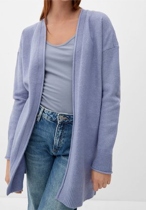 Kvinde iført en lyse lilla åben cardigan over en grå tanktop og blå høj talje jeans mod en hvid baggrund.