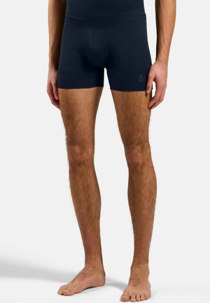 Männliches Model trägt schwarze enganliegende Boxer-Shorts, steht barfuß vor einem schlichten weißen Hintergrund, Unterkörper und Beine sichtbar.