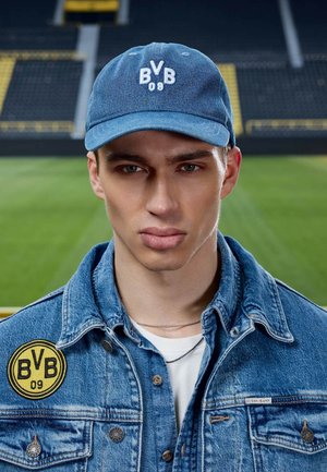 Junger Mann, der eine blaue BVB 09 Kappe und eine Jeansjacke mit BVB 09 Aufnäher trägt, steht in einem Fußballstadion mit verschwommenen Sitzen und Spielfeld im Hintergrund.