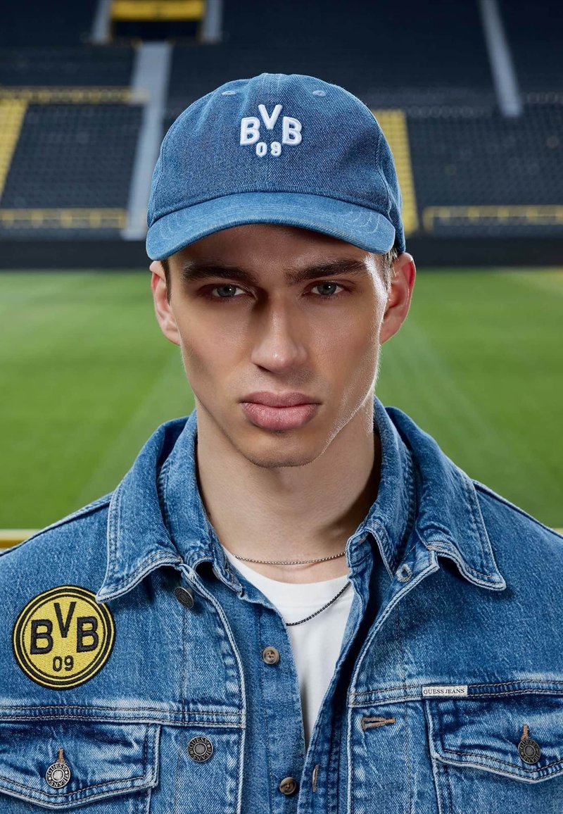 Jeune homme portant une casquette bleue BVB 09 et une veste en jean avec un écusson BVB 09, debout dans un stade de football avec des sièges flous et un terrain en arrière-plan.