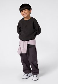 Ragazzo sorridente in felpa nera, pantaloni cargo viola, sneakers bianche, con un maglione rosa chiaro legato in vita, in piedi su uno sfondo neutro.