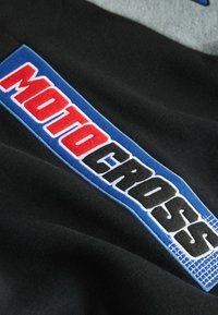Schwarzer Stoff mit einem blauen Patch, auf dem "MOTOCROSS" in roter und schwarzer Stickerei zu sehen ist, vor einem grauen strukturierten Hintergrund präsentiert.