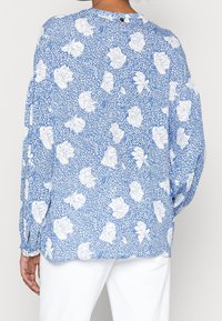 Blusa a maniche lunghe blu con motivo floreale bianco, con una vestibilità leggermente ampia e chiusura a bottoni sulla schiena. Tessuto morbido.