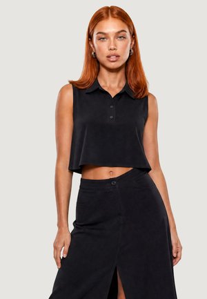 Femme aux cheveux roux portant un haut court noir sans manches avec col et une jupe noire taille haute avec une fente à l'avant, debout devant un fond uni.