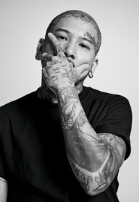 T-shirt noir porté par une personne tatouée, avec des tatouages visibles sur les bras et la tête, comprenant des motifs complexes et des piercings faciaux.