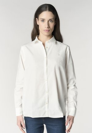 Giovane donna con lunghi capelli castani, che indossa una camicia bianca con bottoni e jeans blu, in piedi davanti a uno sfondo chiaro e semplice.