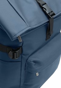 Niebieski plecak z materiału, w którym widoczny jest róg klapy z czarnym zapięciem, regulowanym paskiem oraz prostokątną przednią kieszenią z suwakiem.