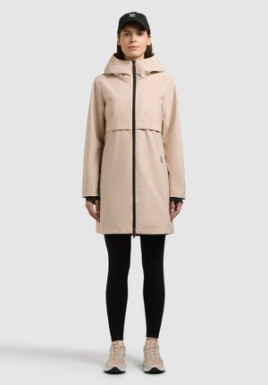 Femeie în picioare, purtând o haină bej cu glugă, colanți negri, adidași bej și o șapcă de baseball neagră pe un fundal simplu.