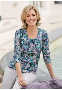 Lächelnde Frau mit kurzem blonden Haar, die eine bunte Blumenbluse und weiße Hosen trägt, sitzend an einem Wasserbecken im Freien.