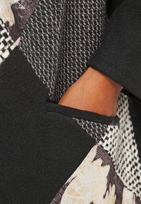Tissu noir avec des patches texturés ; dispose d'un motif tissé en gris et blanc, ainsi qu'un design floral. Comprend une poche latérale visible.