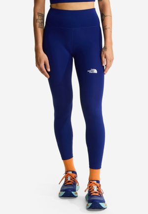Person iført navyblå leggings med The North Face-logo på låret, orange sokker og blå løbesko med orange snørebånd.