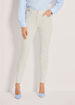 Frau trägt off-white Slim-Fit-Hose mit goldener Kettendetail, kombiniert mit hellblauen High Heels und einer hellblauen Langarmbluse.
