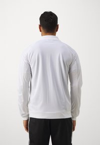 Witte sportieve longsleeve shirt met een hoge kraag, voorzien van een gladde textuur en geribbelde manchetten, gedragen met donkere sportieve broek.