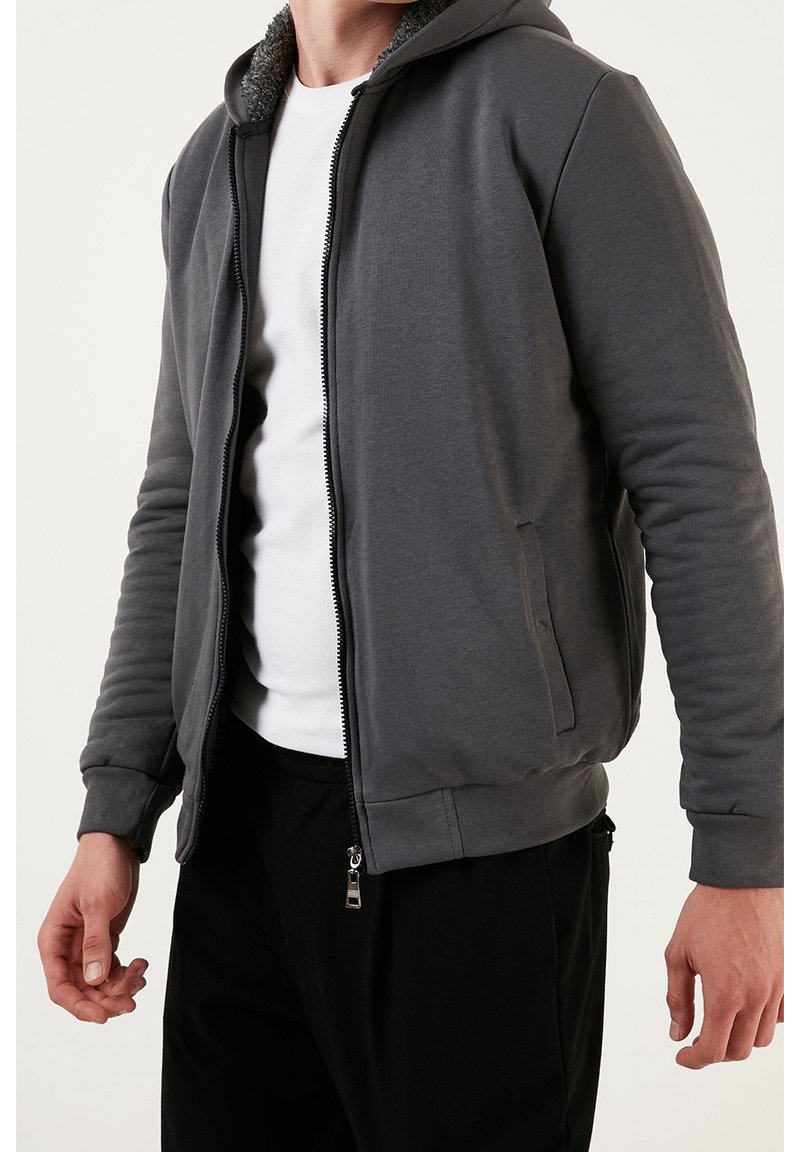 Felpa grigia con zip e cappuccio foderato in pile, polsini e orlo a costine. Abbinata a una t-shirt bianca e pantaloni neri, con tasche laterali.