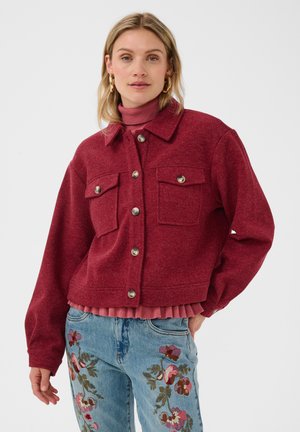 Vrouw draagt een rode jas met knopen over een roze coltrui, gecombineerd met blauwe jeans met kleurrijke bloemenborduursels.