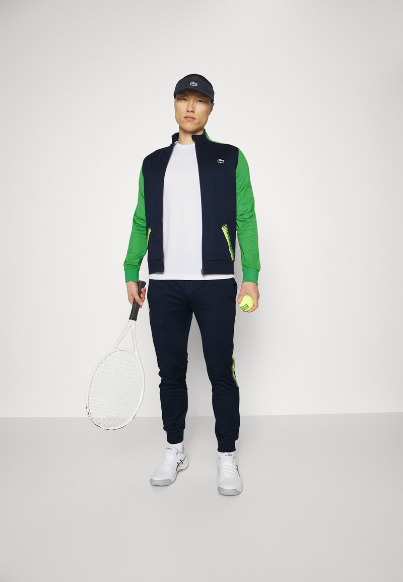 Męski tenisista ubrany w granatowy i zielony dres, białą koszulę, białe trampki i czapkę z daszkiem, trzymający rakietę i żółtą piłkę tenisową.